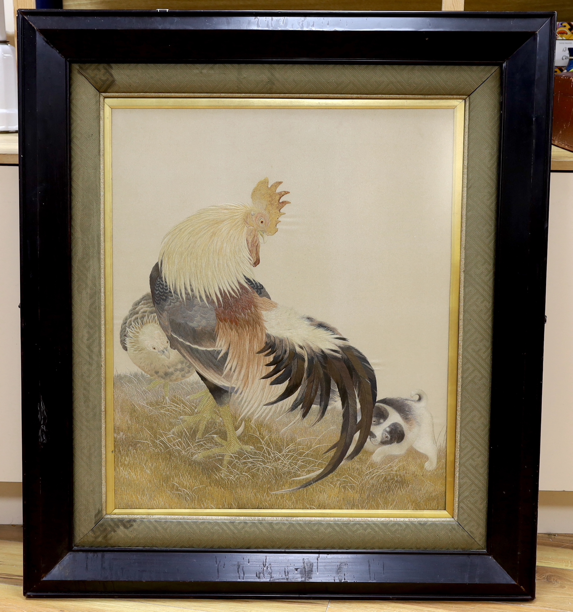A large framed early 20th century Japanese silk polychrome embroidery of a cockerel, it’s chick and puppy by the chicken’s tail feathers, 63cm wide x 75cm high, not including mount or frame
